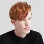 A young person with pale skin and wavy red hair, wearing a black ribbed sweater, looks slightly to the right against a plain light background.