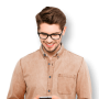 A young man with brown hair and glasses, wearing a light brown button-up shirt, is smiling while looking down at a smartphone he is holding in his hands. The background is plain and neutral.