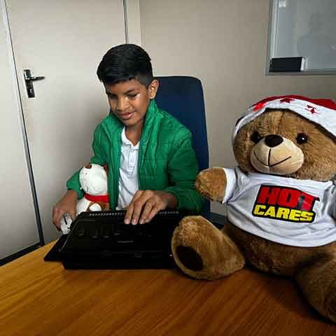 A boy in a green jacket uses a Braille keyboard at a desk, holding a small stuffed dog. Nearby, a large teddy bear wearing a HOT Cares shirt and Santa hat adds Christmas cheer to the scene.