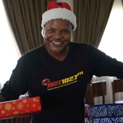 A smiling person wearing a Santa hat and a black Hot 102.7 FM shirt holds two wrapped gifts, supporting the Toy Run, in a room with large windows and curtains.