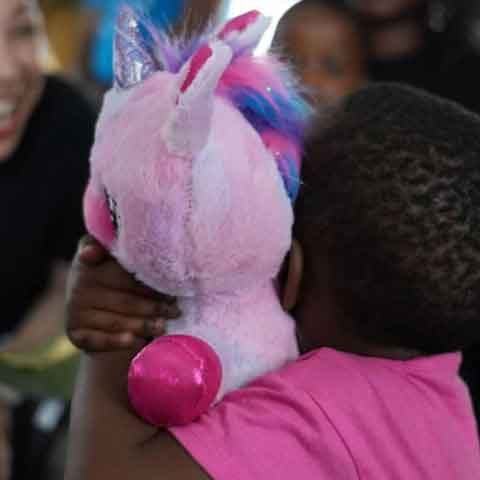 A child in a pink shirt hugs a plush pink unicorn toy, perhaps a favorite find from a recent Toy Run, while an adult nearby smiles warmly in the background.
