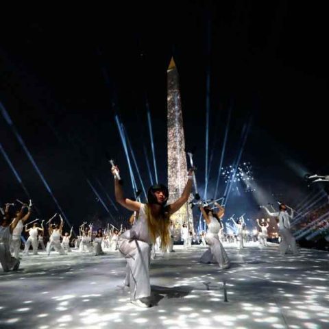 Performers in white costumes dance with dramatic poses and props during a nighttime ceremony, set before a tall illuminated obelisk, as spotlights and beams of light sweep across the scene.