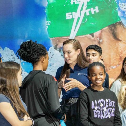 A woman in a blue shirt talks to a group of smiling children and teenagers—A moment to remember with a large image of a swimmer in a green cap and goggles displayed behind them in the pool.