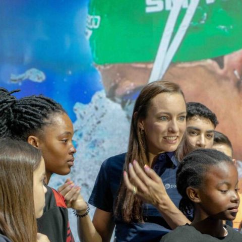 A woman speaks enthusiastically to a group of diverse children, with a large image of a swimmer wearing a green cap and goggles in the background—truly, a moment to remember as everyone appears engaged and attentive.
