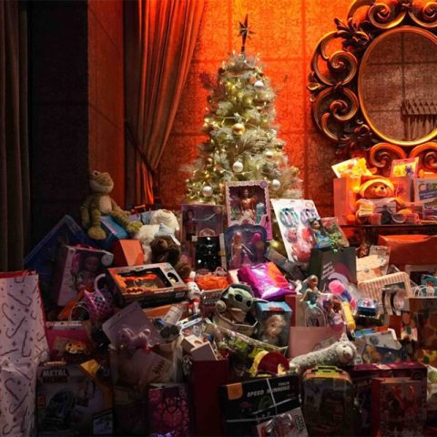 A decorated Christmas tree stands in a warmly lit room beside a gold ornate mirror, surrounded by a large pile of Old Skool wrapped presents and toys, including stuffed animals, dolls, and games.