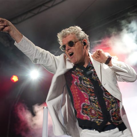 A man in a white suit and sunglasses energetically performs on stage at Marks Park, pointing with one hand and holding a microphone in the other, with dramatic stage lighting and smoke in the background.