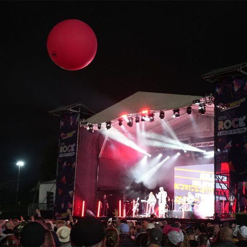 A nighttime outdoor concert at Marks Park shows a crowd facing a brightly lit stage with musicians performing. A large red balloon floats above the audience, and festival banners are displayed on each side of the stage.