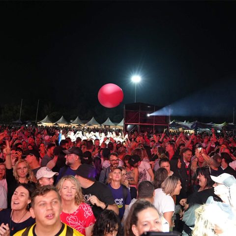 A large crowd enjoys an outdoor nighttime concert at Marks Park, illuminated by stage lights. People are cheering and a giant red balloon floats above the audience, with tents and equipment visible in the background.