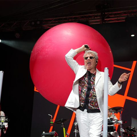 A man in a white suit and patterned shirt sings on stage at Marks Park, holding a microphone with one hand raised. A large pink balloon and colorful stage lights are behind him, with musical equipment and another performer partially visible.