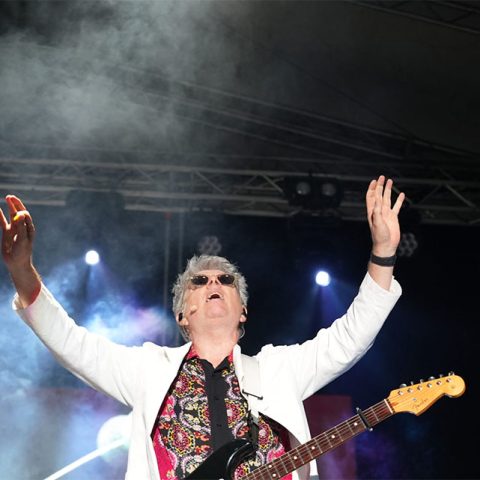 A musician wearing sunglasses, a white jacket, and a patterned shirt plays an electric guitar on stage with arms raised at Marks Park, surrounded by stage lights and smoke effects.