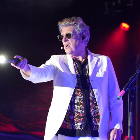 A man in a white suit and sunglasses performs on stage at Marks Park, holding a microphone with a spotlight. The background is dark with red stage lights, and a microphone stand is visible nearby.