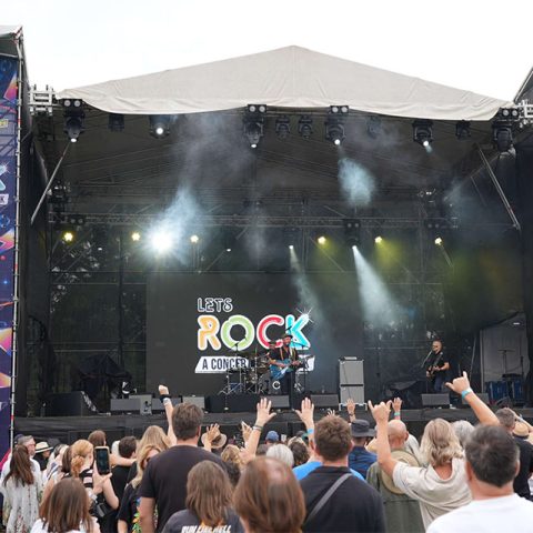 Crowd in front of an outdoor concert stage at Marks Park with Lets Rock: A Concert in the Park displayed on a large screen. Musicians perform under vibrant lights as people in the audience raise their hands and enjoy the show.