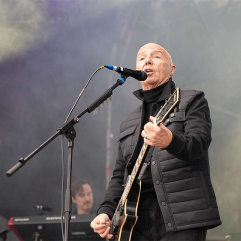 A bald man in a black jacket sings into a microphone while playing electric guitar on stage at Marks Park. In the background, amid stage lights and fog, a keyboard player is also visible.