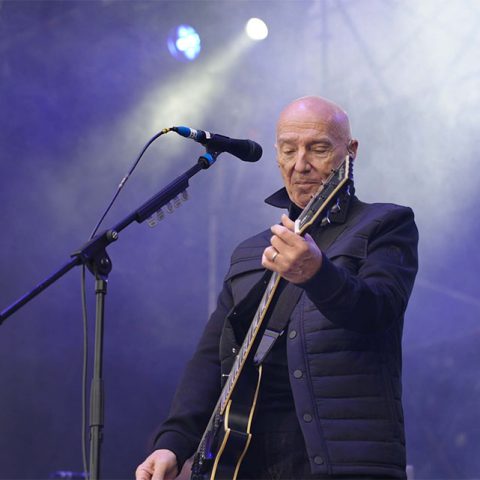 A bald man wearing a dark jacket plays an electric guitar and sings into a microphone on the Marks Park outdoor stage, surrounded by blue and white stage lights and mist.