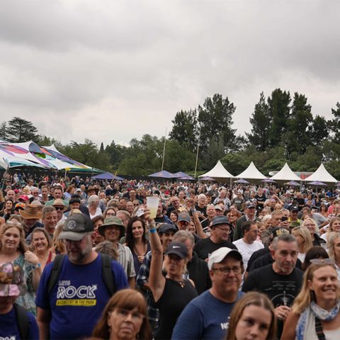 A large crowd of people stands outdoors at Marks Park for a festival or concert, some smiling and wearing hats. Colorful tents and white marquees are visible in the background, with trees and a cloudy sky overhead.