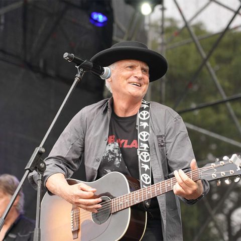 A man wearing a black hat and shirt plays an acoustic guitar and sings into a microphone on an outdoor stage at Marks Park, with another musician visible in the background.
