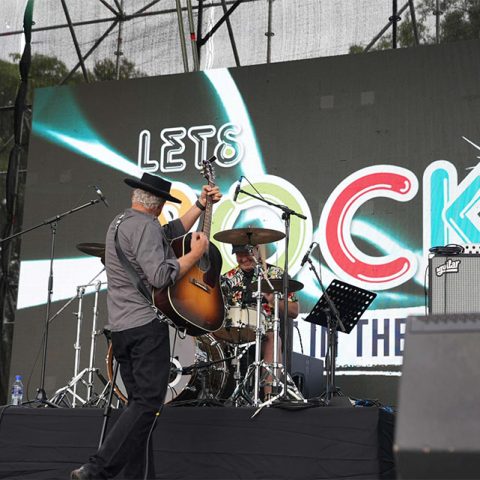 Two musicians perform onstage at Marks Park; one plays electric guitar, the other drums. Behind them, a large screen displays LETS ROCK in colorful letters, with visible stage equipment and lighting set outdoors.