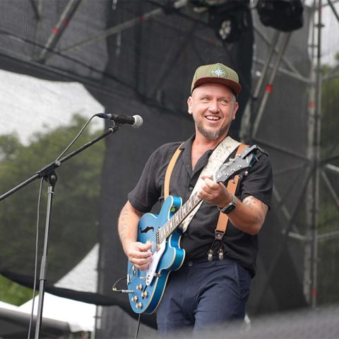 A smiling man wearing a cap plays a blue electric guitar and sings into a microphone on an outdoor stage at Marks Park, with scaffolding and trees in the background.