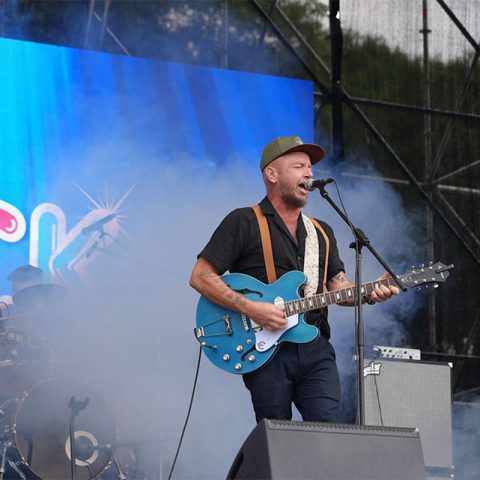 A musician in a cap sings and plays a blue electric guitar on an outdoor stage at Marks Park, surrounded by stage smoke. A drummer is partially visible in the background, and colorful lights illuminate the scene.