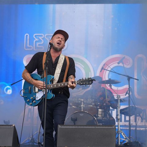 A male musician wearing a cap and black shirt passionately sings and plays an electric guitar on stage at Marks Park, with a drummer behind him. A colorful Lets Rock sign is visible in the background.