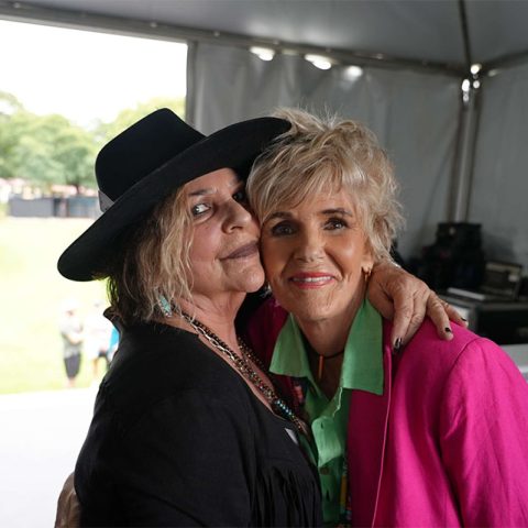 Two older women smiling and embracing under a tent at Marks Park. One wears a black hat and dark clothes; the other sports a bright pink blazer and green shirt. Outdoor setting with trees visible in the background.