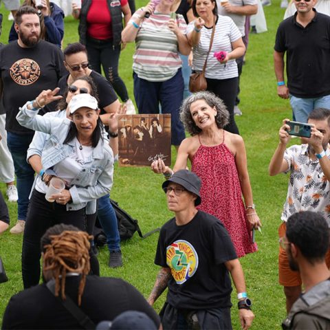 A diverse group of people enjoy an outdoor event on the grass at Marks Park. Some are smiling, holding drinks, taking photos, or posing; one person holds a sepia photo. The mood is lively and festive.