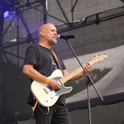 A bald man wearing a black t-shirt plays an electric guitar and sings into a microphone on an outdoor stage at Marks Park, with stage lights and equipment visible behind him.