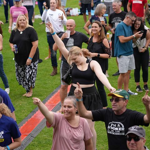 A crowd of people enjoy an outdoor event on the green grass of Marks Park. Some are smiling, dancing, and raising their arms. Most are casually dressed, and a few hold drinks. The atmosphere is lively and festive.
