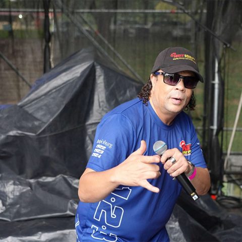 A man wearing sunglasses, a cap, and a blue T-shirt holds a microphone and gestures with his hand while performing on an outdoor stage covered with tarps at Marks Park.
