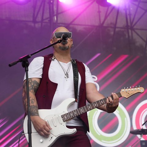 A bald man wearing sunglasses and a red vest plays an electric guitar and sings into a microphone on stage at Marks Park, with colorful lights and a vibrant background behind him.