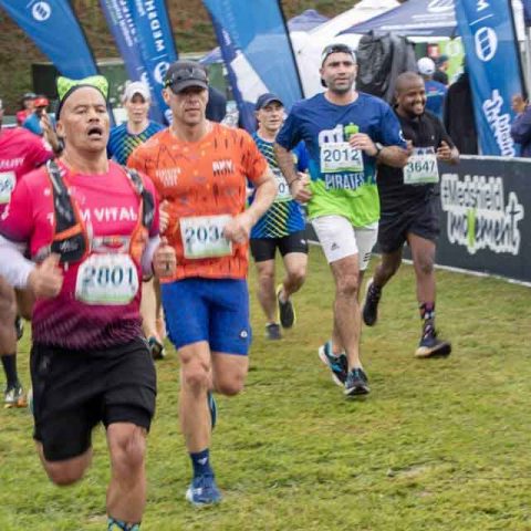 A group of runners wearing race bibs approach the finish line at an outdoor running event in Marks Park, with banners and spectators visible in the background on a grassy field.