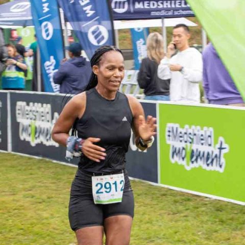 A woman wearing a black athletic outfit and bib number 2912 runs past a green MedShield banner at the Marks Park outdoor race event, with onlookers and other runners in the background.