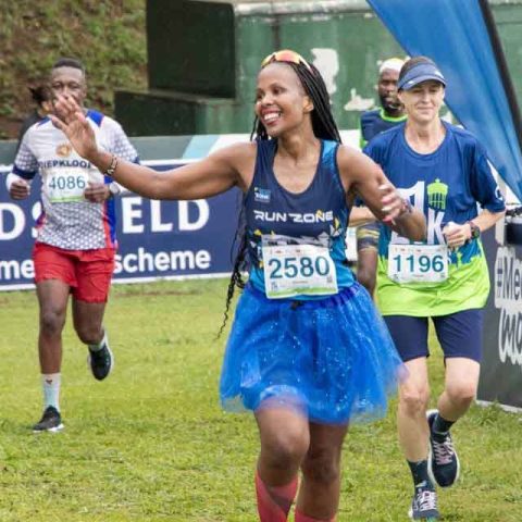 A smiling woman in a blue tutu, race bib 2580, runs toward the finish line with arms outstretched at Marks Park. Other runners follow behind her on a grassy field during a race event.