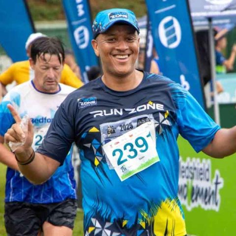 A smiling runner wearing a blue Run Zone shirt and cap, with race bib 239, gestures with both index fingers raised as he crosses the Marks Park race finish line. Other participants and banners are visible in the background.