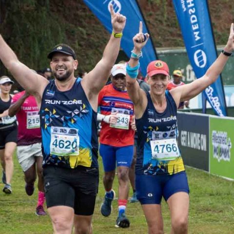 Runners in athletic gear smile and raise their arms in victory as they approach the finish line at Marks Park, surrounded by other participants and event banners.