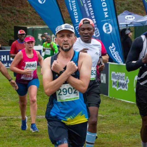A group of marathon runners near the finish line at Marks Park; one runner at the front, wearing bib number 2902, crosses his arms over his chest while others focus ahead. Event banners and spectators are visible in the background.