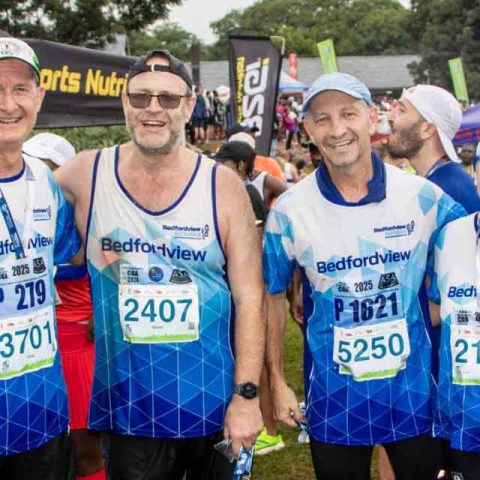 Four smiling runners in matching Bedfordview marathon shirts and race bibs pose together outdoors at a Marks Park race event, surrounded by other participants and colorful banners.