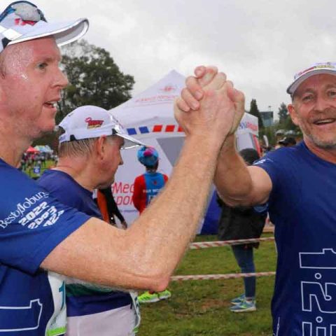 Two male runners in blue shirts and race bibs clasp hands and smile in celebration at an outdoor event in Marks Park, with other participants and a white tent visible in the background.