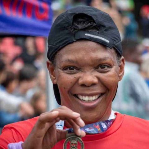 A smiling person wearing a red shirt and a backwards black cap holds up a medal at Marks Park, with a crowd blurred in the background, suggesting a race or marathon event.