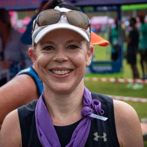 A smiling woman wearing a white cap, sunglasses, and a purple scarf holds up a medal at Marks Park during an outdoor event. People and tents are visible in the blurred background.