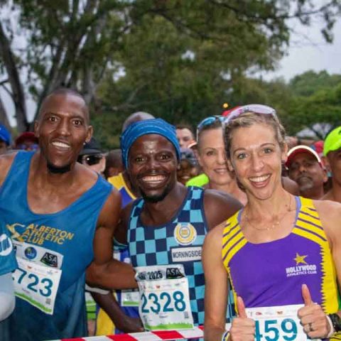 A group of marathon runners, men and women, stand smiling at the starting line in Marks Park outdoors, wearing colorful race bibs and athletic gear, with trees and other participants visible in the background.