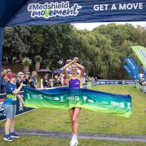 A runner in athletic gear crosses the finish line at Marks Park, breaking the tape with arms raised in victory at an outdoor race event. Spectators and officials cheer under a “Medshield Movement” banner.