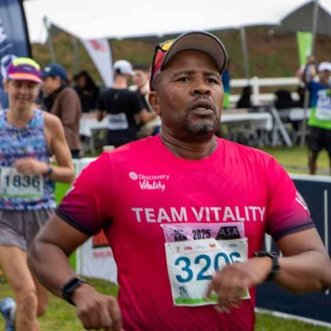 A man wearing a red Team Vitality shirt and race number 3206 runs in a race at Marks Park, with another runner and event tents visible in the background.