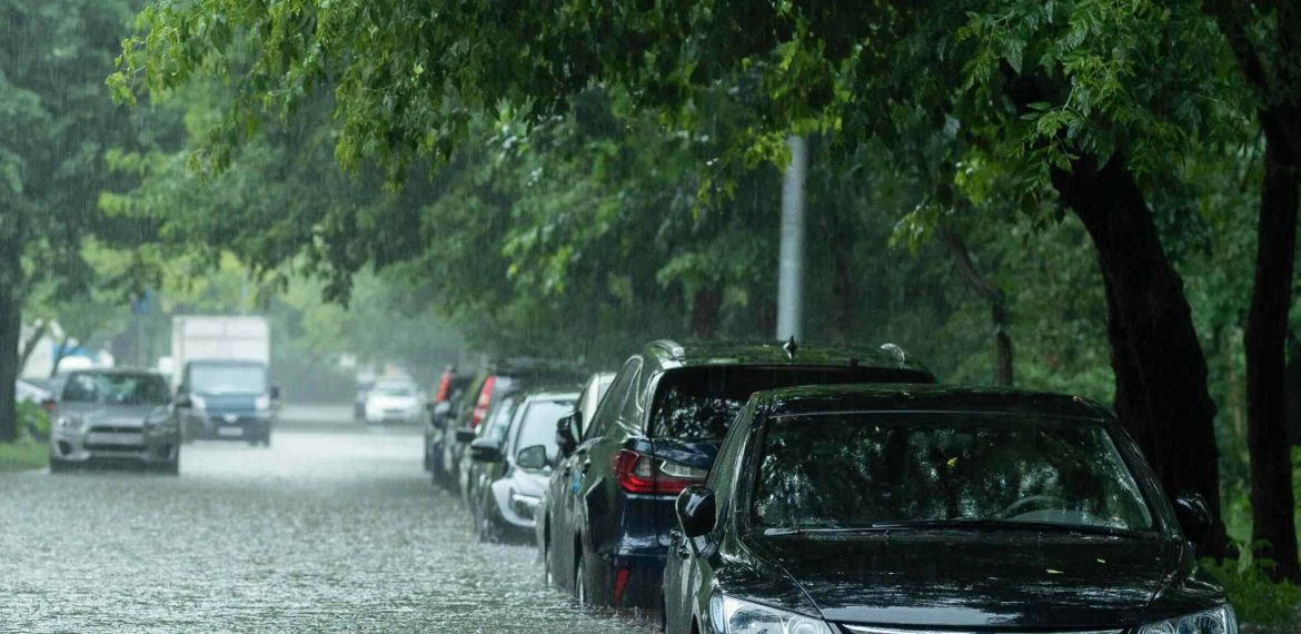 Cars are parked on a street flooded with rainwater, with water reaching up to the lower doors; trees line the street as heavy rain from severe weather continues to fall, creating a gray and gloomy atmosphere.
