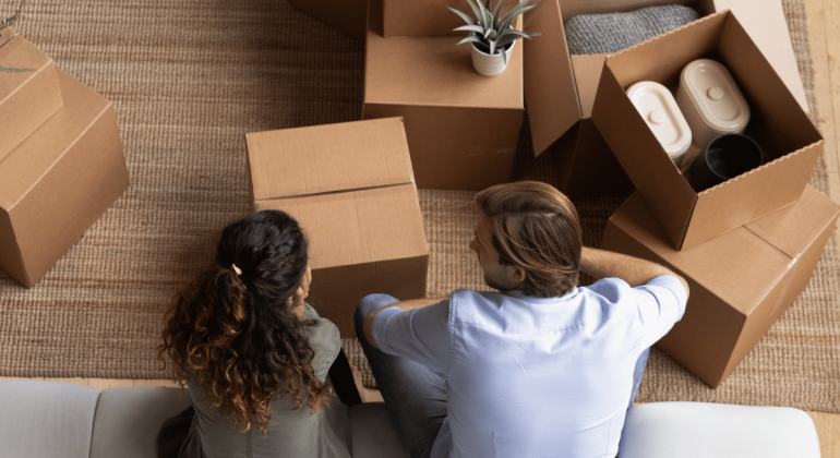 Two homeowners sit on a white couch, seen from above, surrounded by open cardboard boxes with household items inside on a woven rug, suggesting they are moving in or unpacking.
