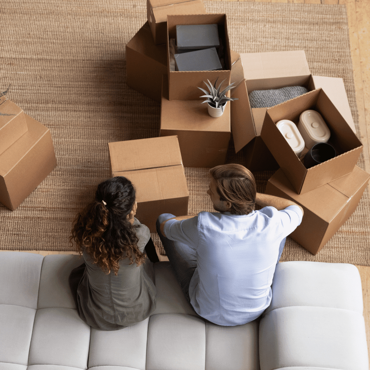 Two homeowners sit on a white couch, seen from above, surrounded by open cardboard boxes with household items inside on a woven rug, suggesting they are moving in or unpacking.