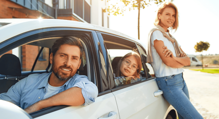 A smiling man sits in the driver’s seat of a white car, with a young girl leaning out of the back window. Her parents stand nearby, all appearing happy in front of a modern house on a sunny day.