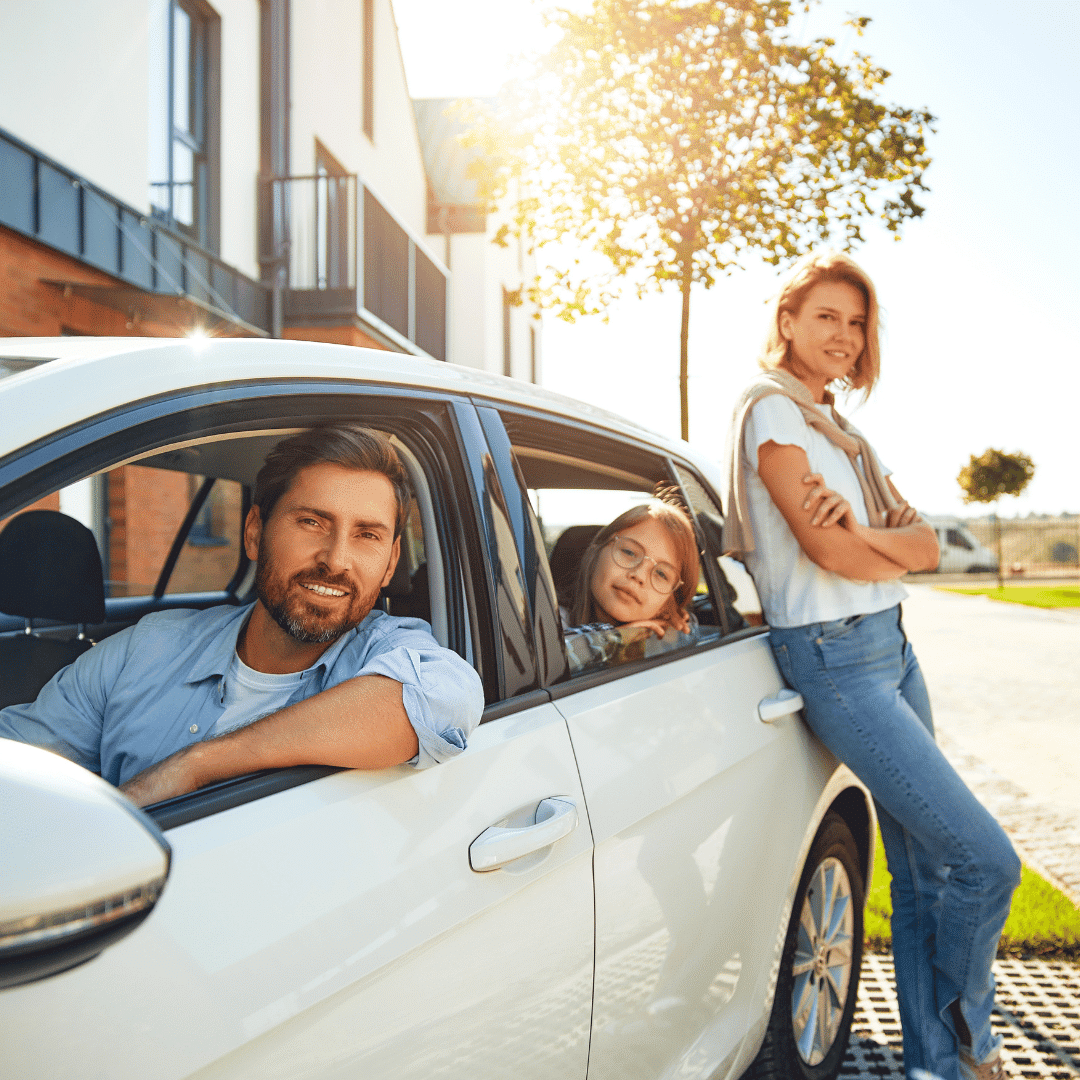 A smiling man sits in the driver’s seat of a white car, with a young girl leaning out of the back window. Her parents stand nearby, all appearing happy in front of a modern house on a sunny day.