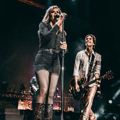A female singer in a sparkly outfit and patterned boots sings into a microphone, while a guitarist—reminiscent of Roxette band members—smiles beside her on a stage with dramatic lighting.