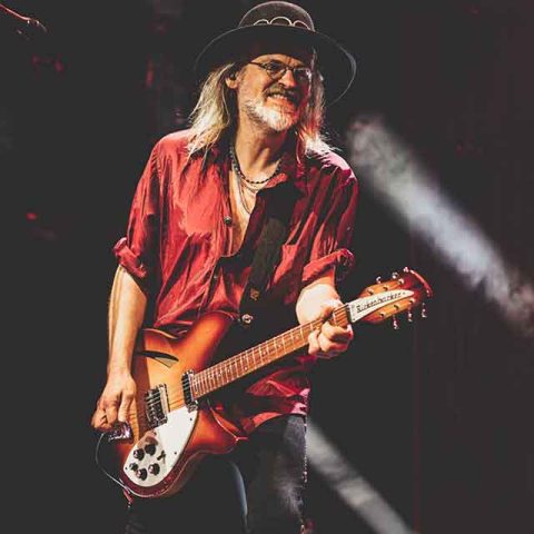 A musician with long hair, sunglasses, and a black hat plays an electric guitar on stage. Reminiscent of Roxette band members, he wears a red shirt and smiles under vibrant stage lights in a lively concert setting.